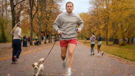 A man in a grey sweatshirt and red shorts jogs on a park path with a small dog on a leash, surrounded by other people exercising in an autumn park.