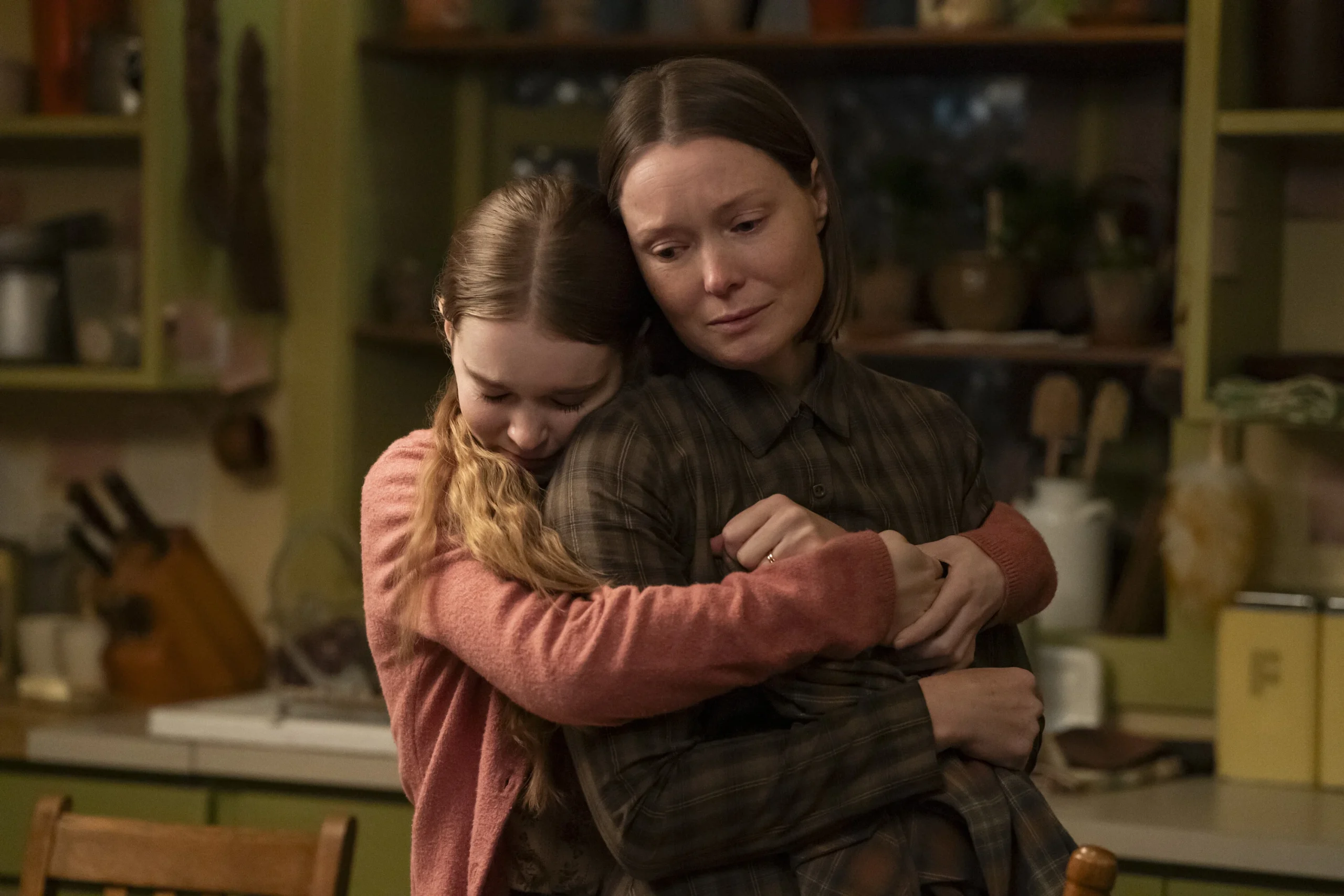 A younger person in a pink sweater hugs an older woman in a brown plaid shirt from behind in a kitchen.
