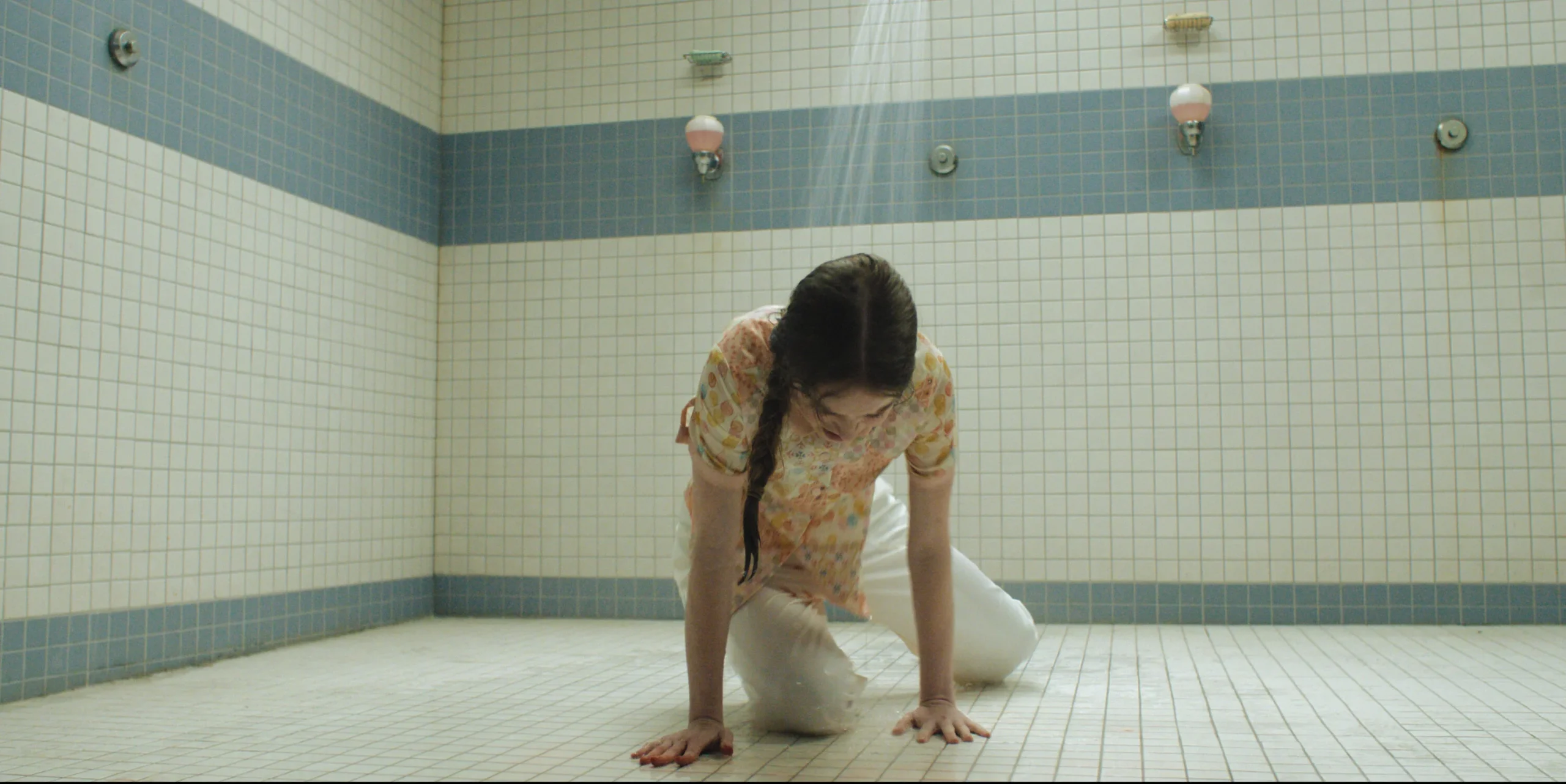 A person with a braided ponytail kneeling under a shower in a tiled communal shower room.
