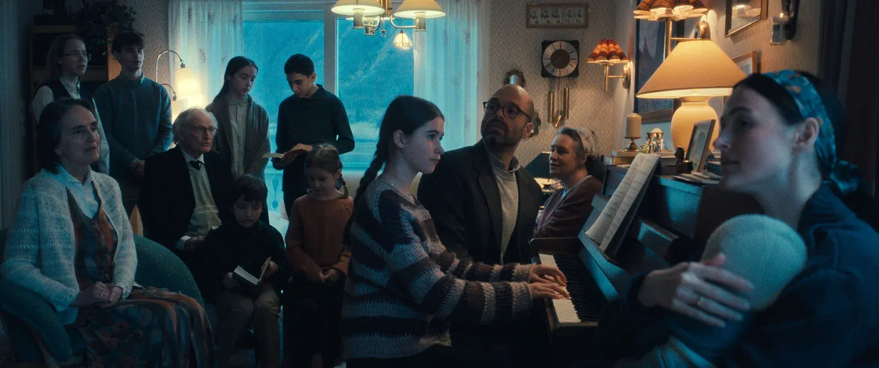 A group of people gathered in a living room, some reading books, while a young girl plays the piano for them.