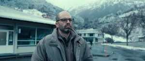 A bald man with glasses and a beard wearing a grey jacket, standing outdoors in a mountainous, snowy setting.