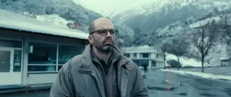 A bald man with glasses and a beard wearing a grey jacket, standing outdoors in a mountainous, snowy setting.