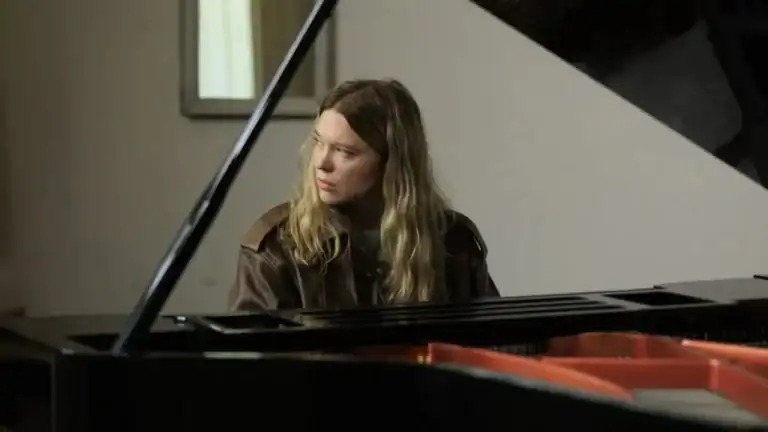 A woman with long blonde hair sits at a grand piano looking off to the side.