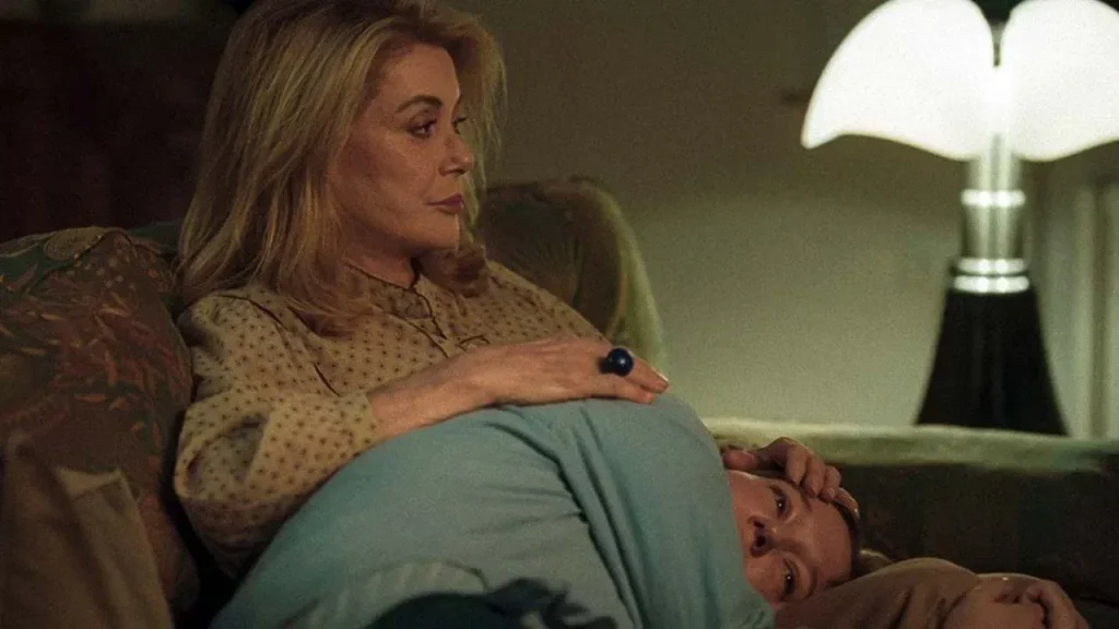 A woman sits on a sofa, resting her hand on the head of another person who is lying in her lap.