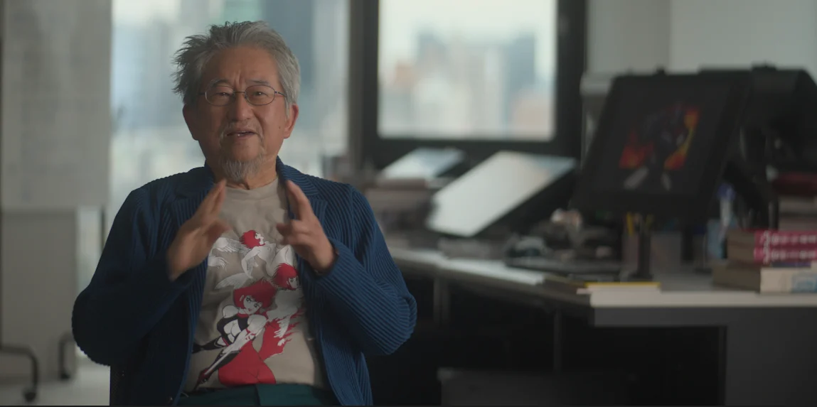 An older man with gray hair and glasses wearing a patterned t-shirt and blue cardigan sits in an office, gesturing with his hands.