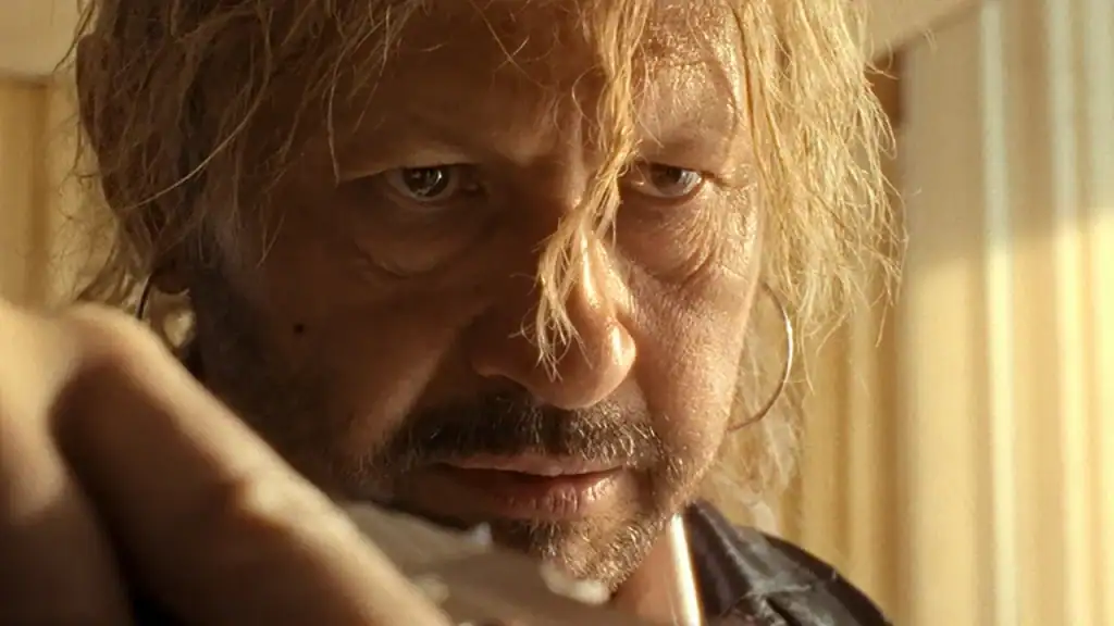 Close-up of a middle-aged man with messy blonde hair and hoop earrings, looking intensely toward the camera.