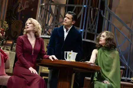 Two women in elegant dresses and a man in a velvet blazer stand around a table on a stage set.