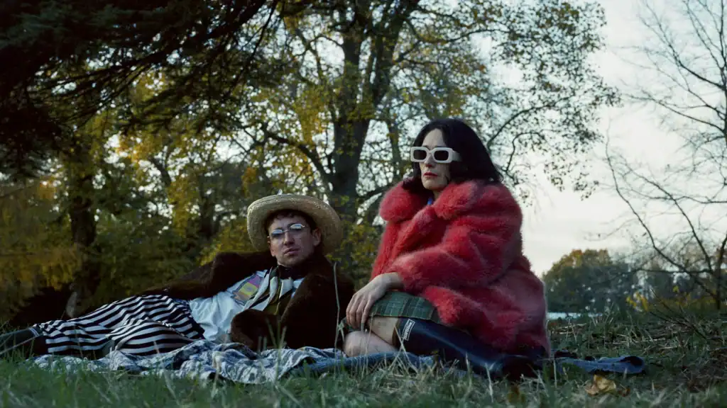 Two people sitting on a grassy field; one person wears a straw hat and striped pants, and the other wears a red faux-fur coat and white-rimmed sunglasses.