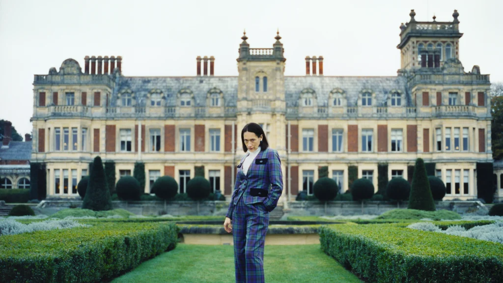 A person wearing a plaid suit stands in a formal garden in front of a large stone manor house.