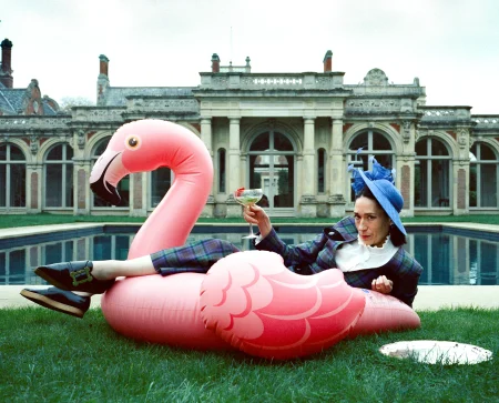 A woman wearing a plaid suit and blue fascinator reclines on a large inflatable pink flamingo beside a swimming pool, holding a cocktail glass.