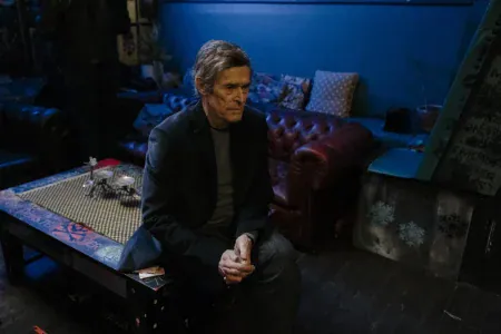 Willem Dafoe sitting on a patterned table in a dimly lit room with blue walls and a red leather sofa.