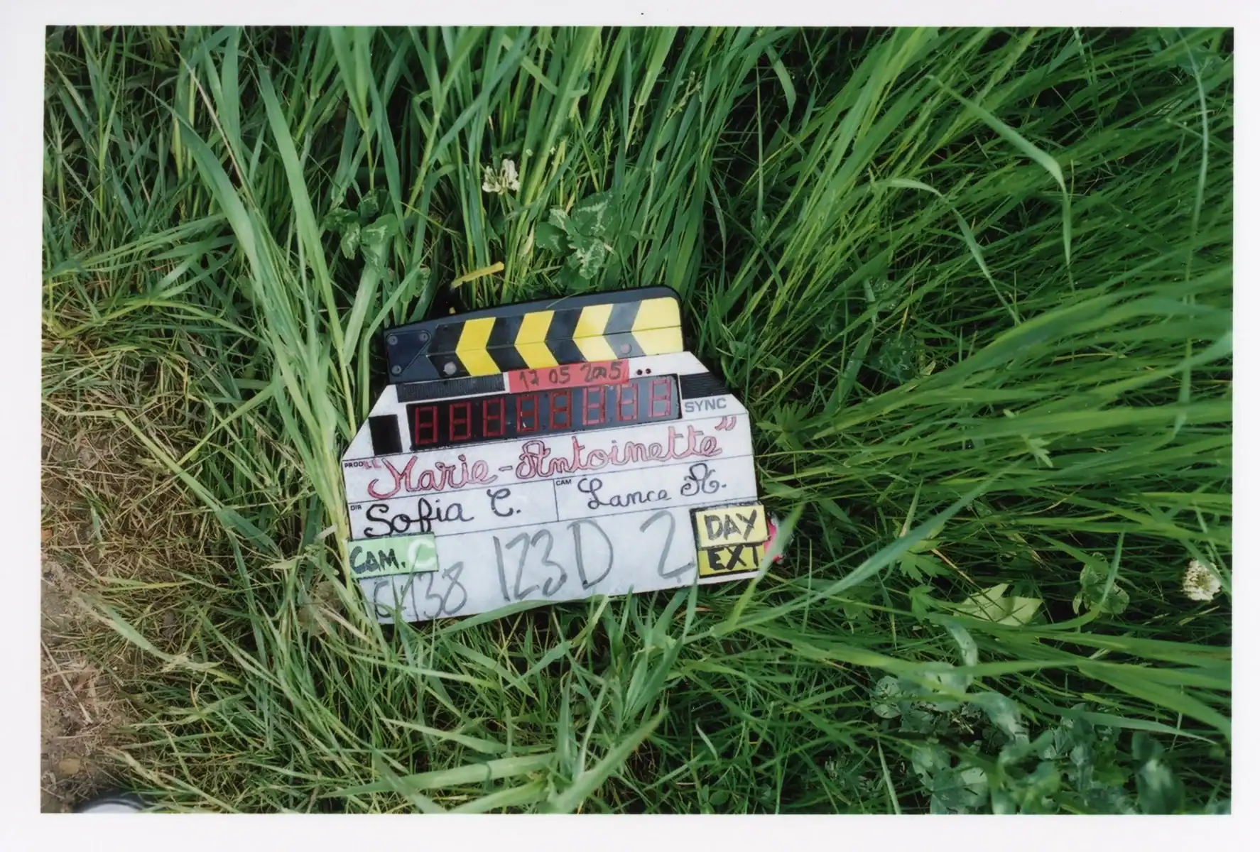 A film clapperboard resting in tall grass, labeled with the production “Marie-Antoinette,” director “Sofia C.”, and cinematographer “Lance A.”
