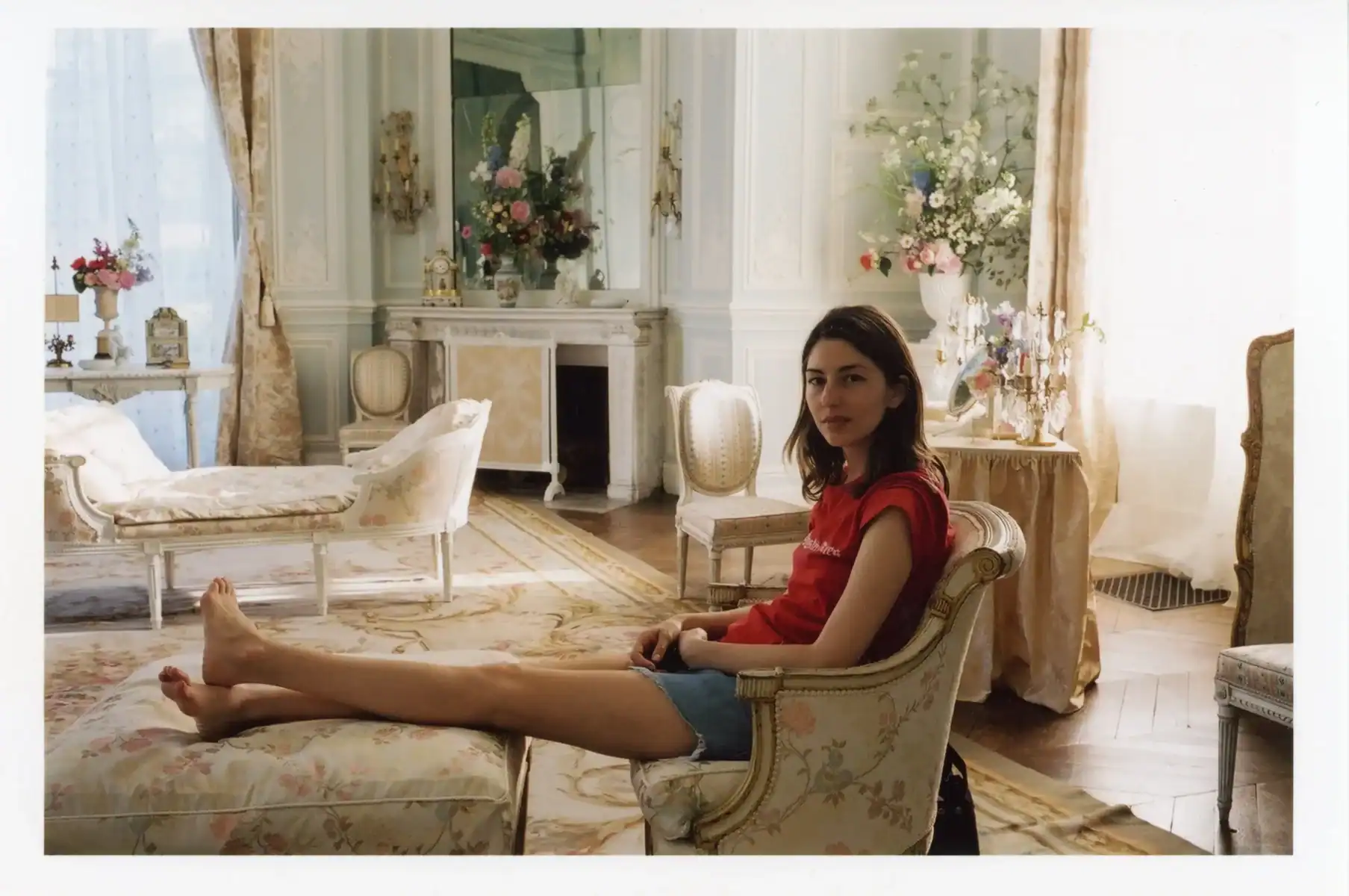 A woman in a red shirt and denim shorts sitting in an armchair with her feet resting on an ottoman in a formal, light-filled room.
