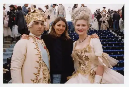 ‘Making Marie Antoinette’ Review – Behind-The-Scenes Documentary Is A Moving Portrait Of A Bond Between A Mother And Daughter Sofia Coppola stands between two actors dressed in elaborate 18th-century French court attire on a staircase decorated with fleur-de-lis patterns.