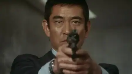 Close-up of a serious-looking man in a dark suit aiming a handgun directly toward the viewer with both hands.