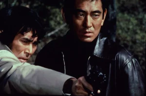 Two men, one in a light jacket and one in a black leather jacket, looking intensely while one holds a handgun.