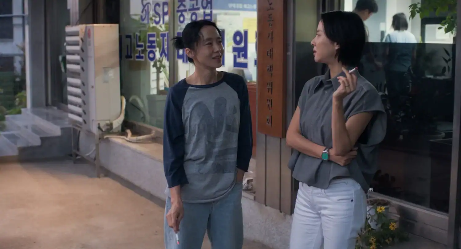 Two women stand outside a storefront, talking and holding cigarettes.