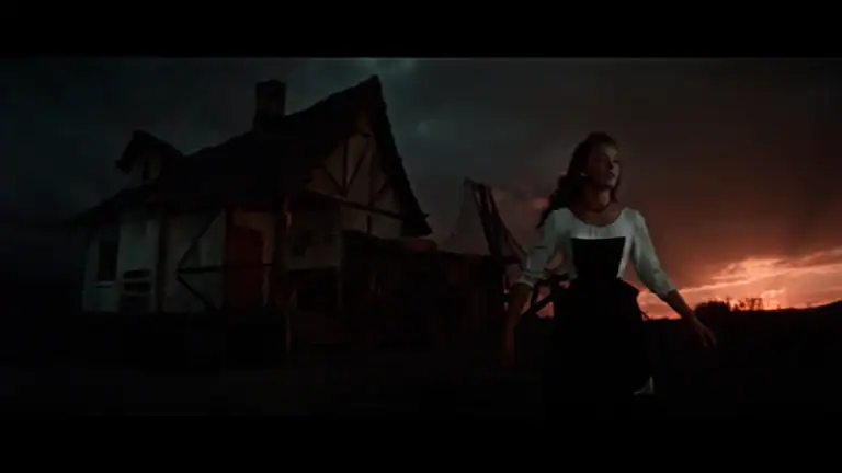 A woman in a white blouse and dark bodice stands in front of a rustic house at sunset.