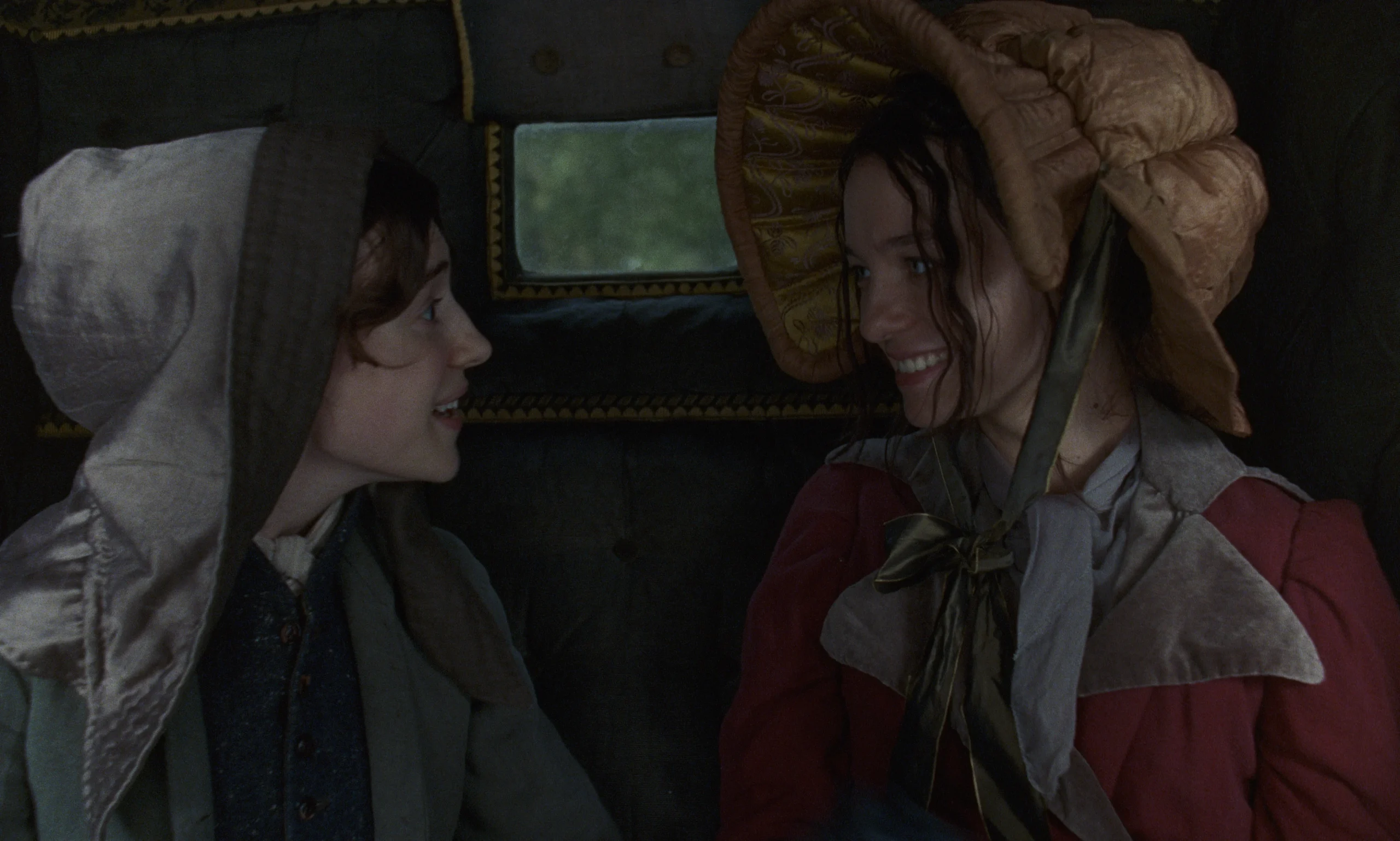 Two women wearing historical bonnets sit inside a carriage, looking at each other.