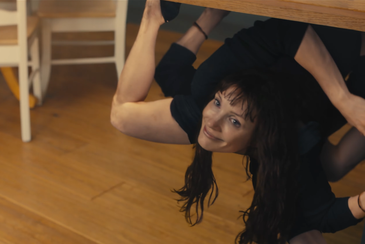 A woman with long dark hair contorted beneath a wooden table, looking up toward the camera.