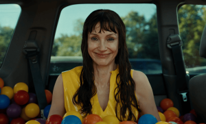 A woman in a yellow top smiles while sitting in the trunk of a car filled with colorful plastic balls.