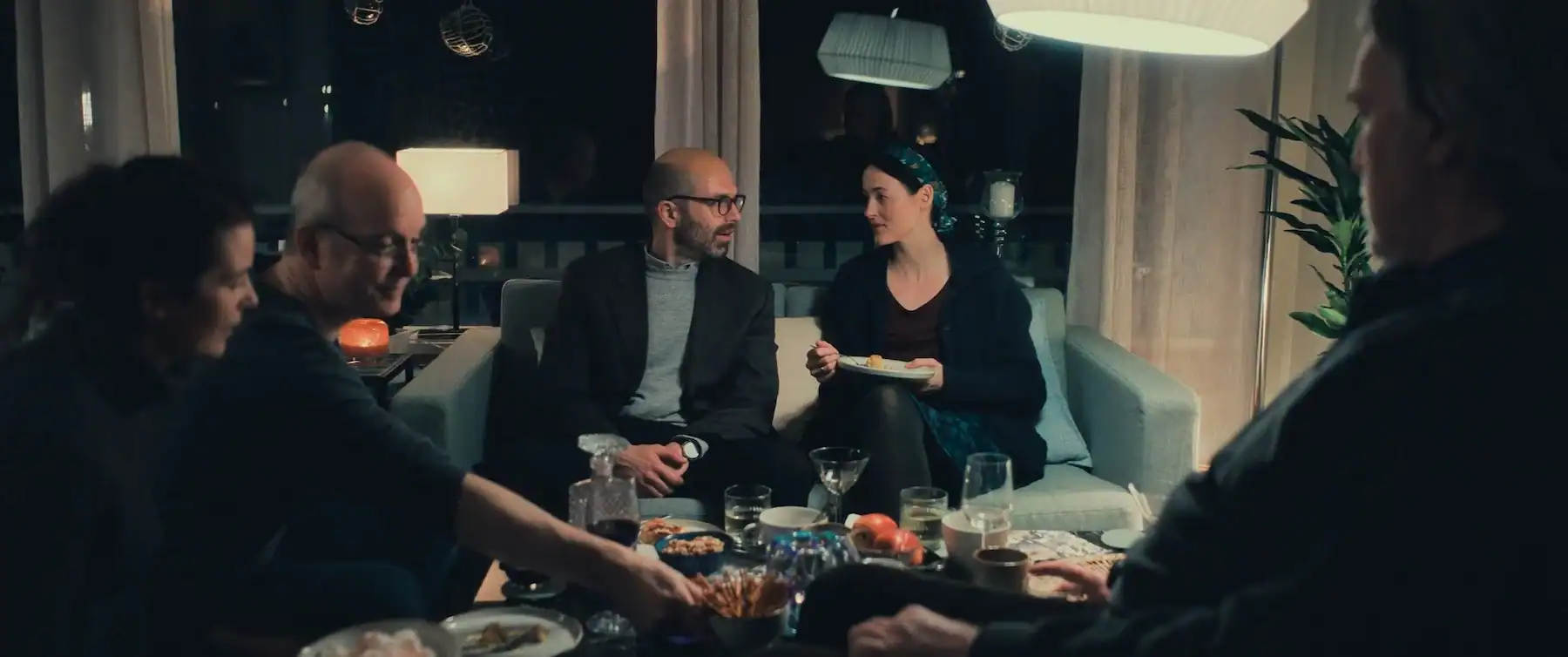 A group of five people sitting in a living room around a coffee table filled with food and drinks.
