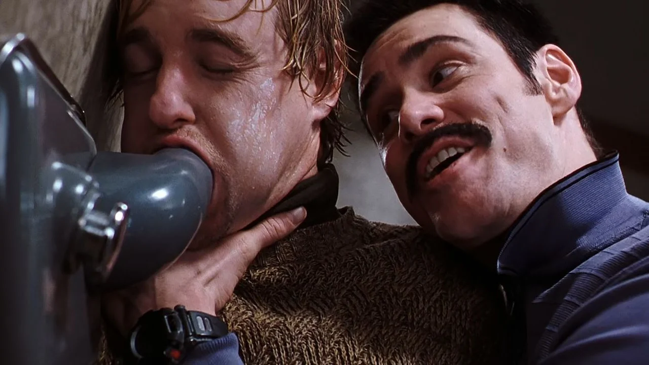 A man with a mustache holds another man by the neck, pushing his face toward a drinking fountain.