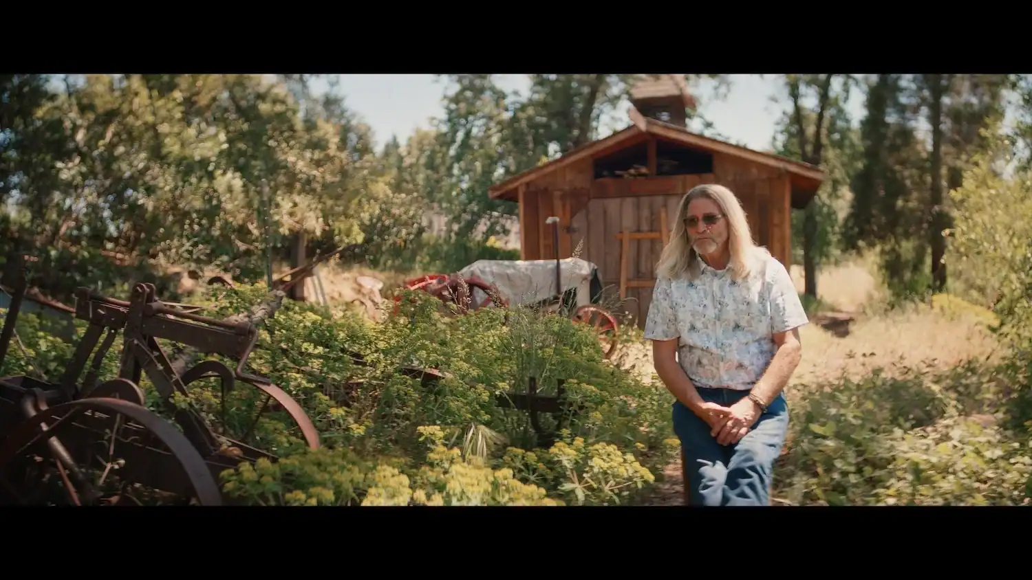 An older man with long gray hair and sunglasses sits in a garden in front of a small wooden shed and old farm equipment.