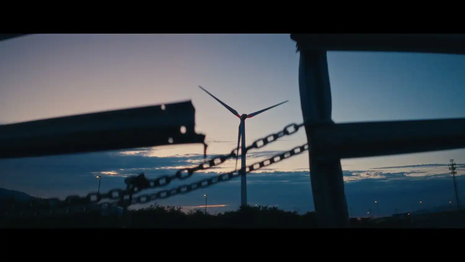 A wind turbine viewed at dusk through a gap in a chain-link fence.