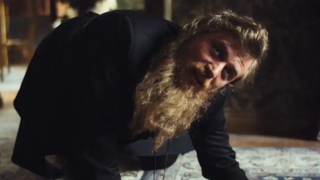 A man with a long beard wearing a black jacket crouches on the floor, looking upward.