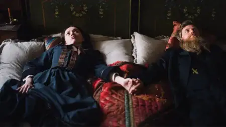A woman in a blue dress and a bearded man in a dark coat lie side by side on a bed, holding hands and looking upward.