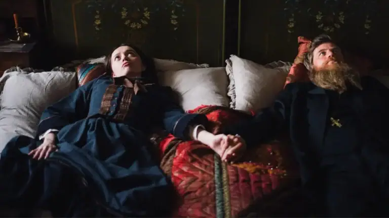 A woman in a blue dress and a bearded man in a dark coat lie side by side on a bed, holding hands and looking upward.