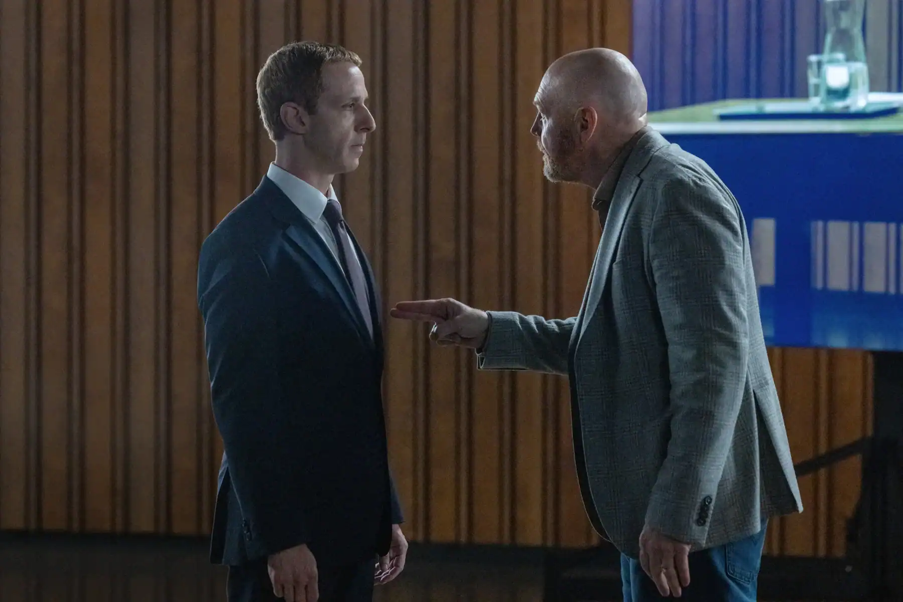 A man in a suit and tie stands still while another man in a plaid blazer gestures toward him with a pointed finger.