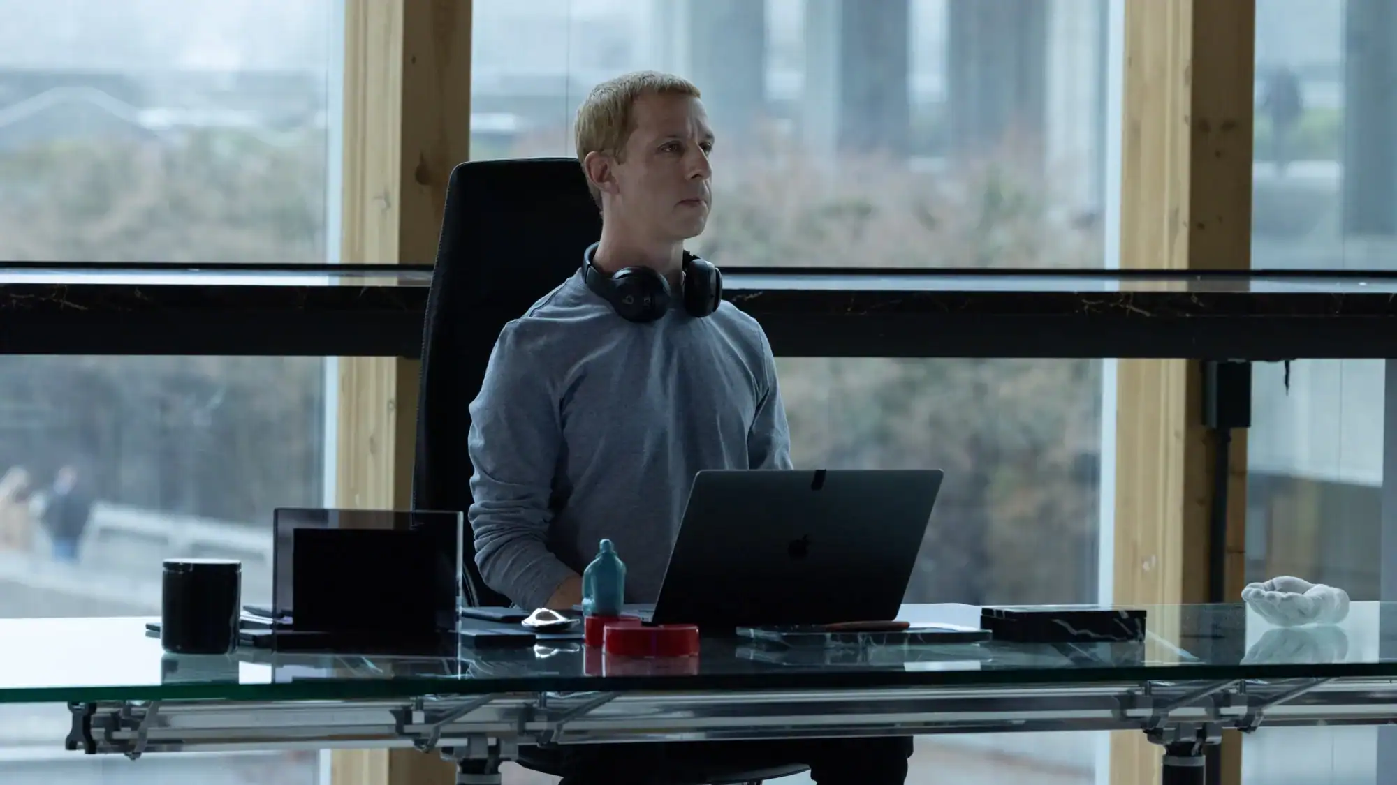 A man sits at a glass desk in front of a laptop with headphones around his neck, facing a large window.