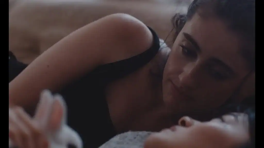 A dark-haired woman in a black sleeveless top lies on her side, gazing at another person whose face is partly visible in the foreground.