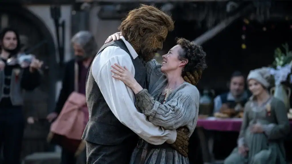 A woman in a rustic dress embraces a man with a textured, mask-like face and a beard, while people in period clothing look on in the background.