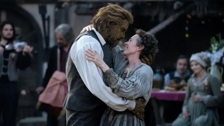 A woman in a rustic dress embraces a man with a textured, mask-like face and a beard, while people in period clothing look on in the background.