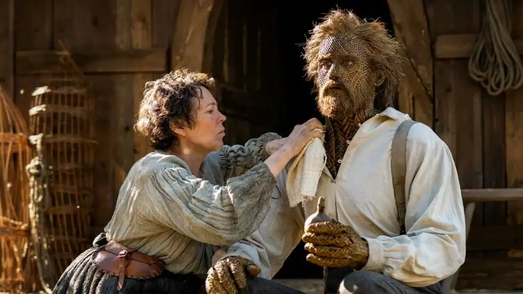 A woman wipes the face of a man with skin made of textured, woven material.