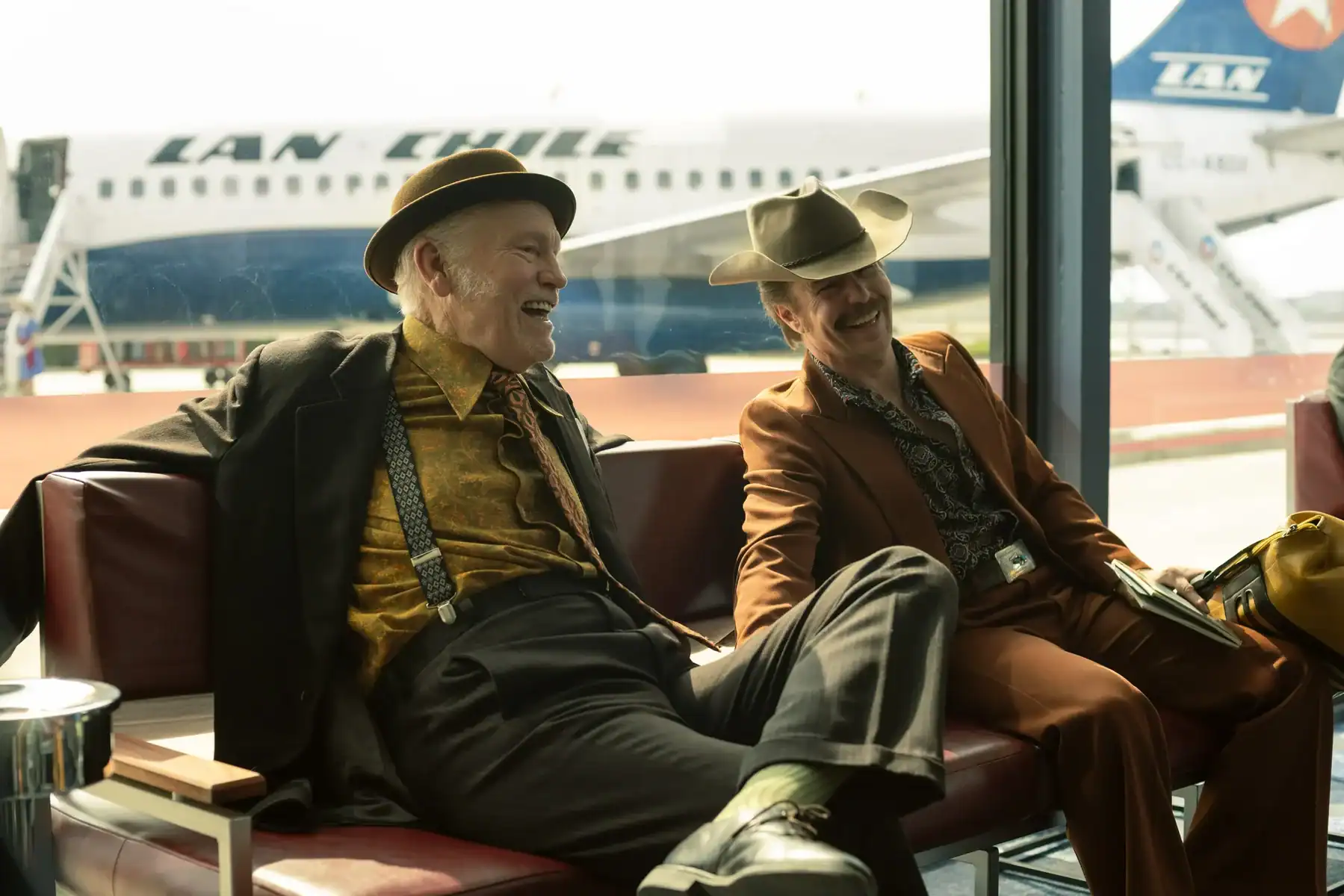 Two men in vintage suits and hats laughing while seated in an airport terminal in front of a LAN Chile airplane.