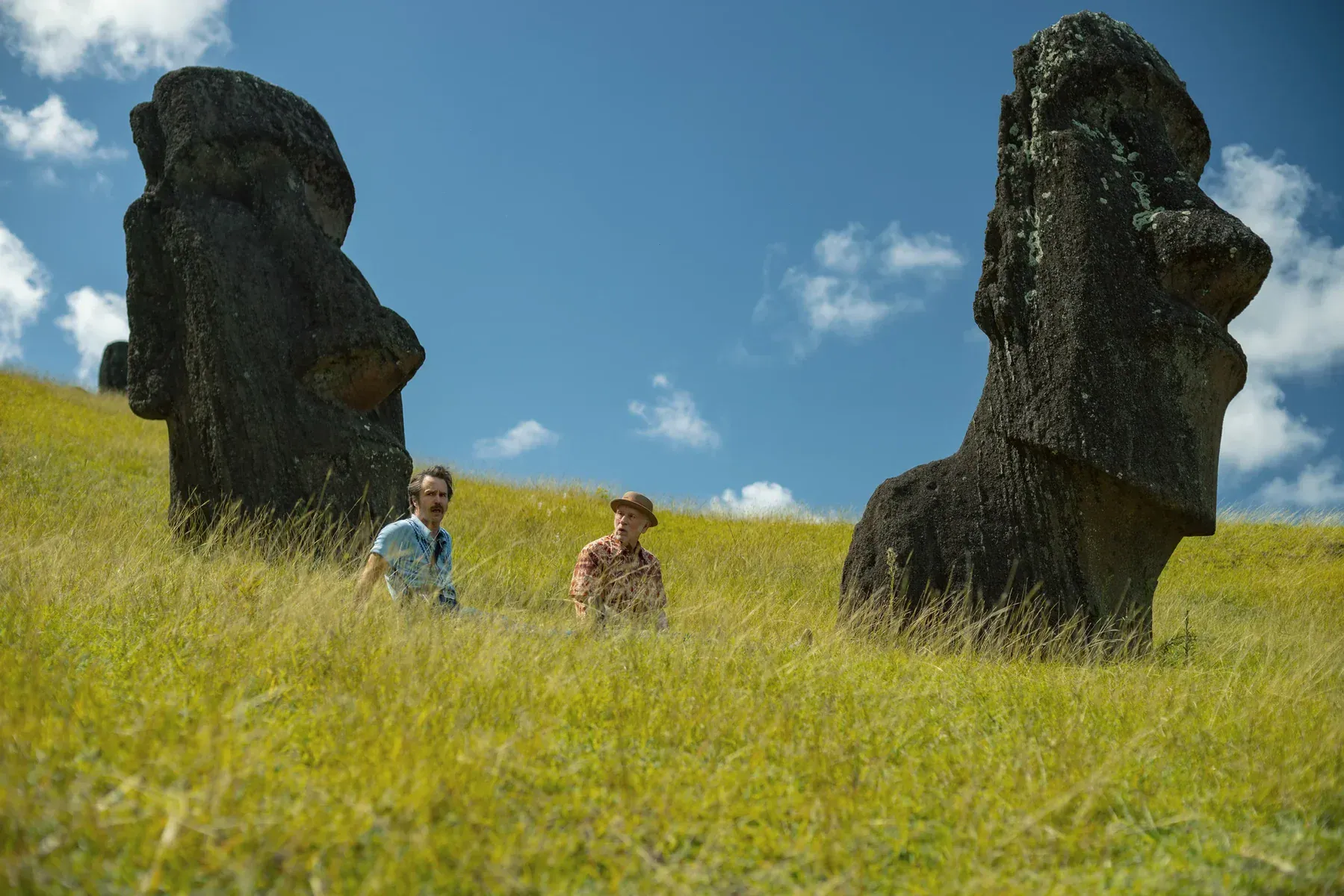 Two men sit in a grassy field between two large stone moai statues on Easter Island.