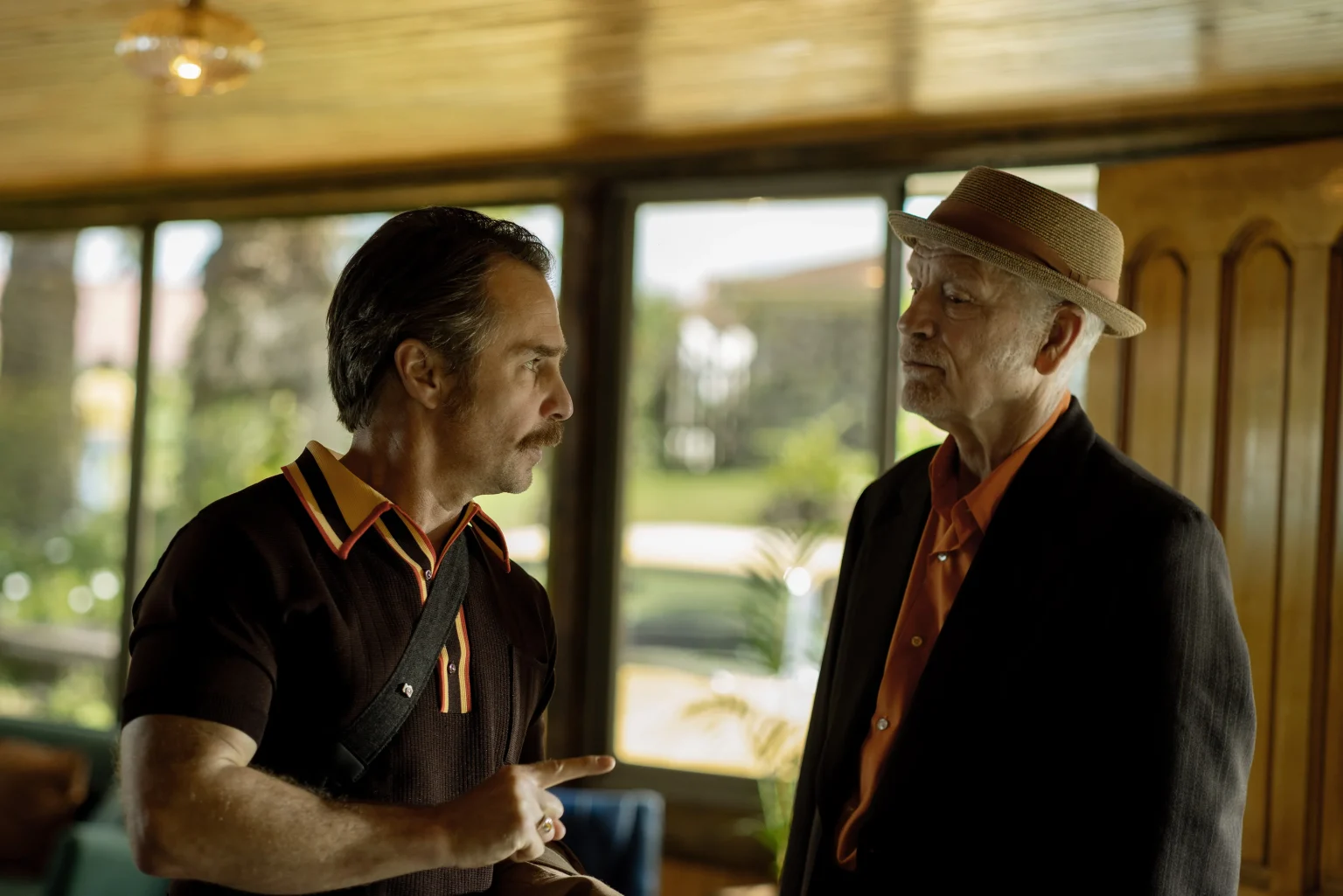 A man with a mustache gesturing while speaking to an older man wearing a straw fedora and a dark blazer.