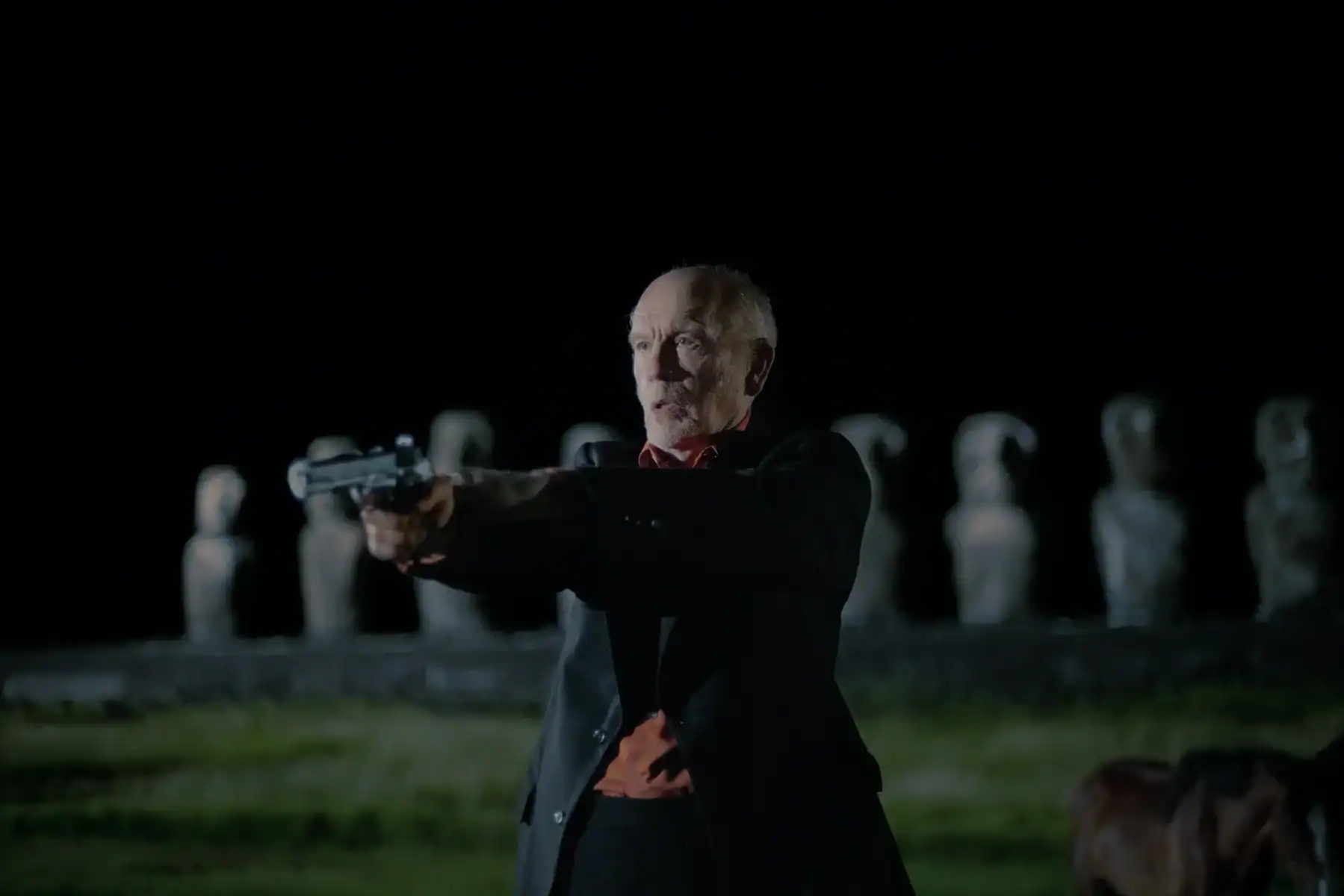 An older man in a suit points a handgun forward at night, with stone statues in the background.
