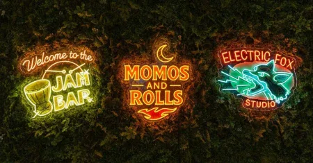Three neon signs mounted on a leafy green wall reading “Welcome to the Jam Bar,” “Momos and Rolls,” and “Electric Fox Studio.”