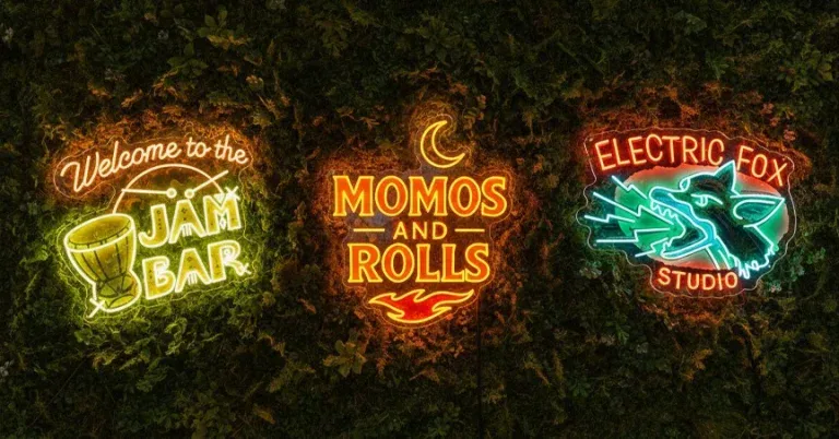 Three neon signs mounted on a leafy green wall reading “Welcome to the Jam Bar,” “Momos and Rolls,” and “Electric Fox Studio.”