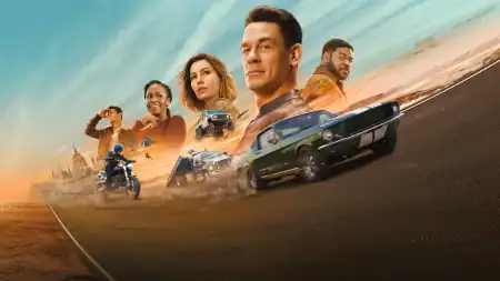 Movie poster featuring John Cena and the cast above a green muscle car, a motorcycle, and a truck on a desert road with the Budapest Parliament building in the distance.
