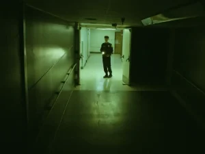 A security guard holding a flashlight stands in a dimly lit hallway.