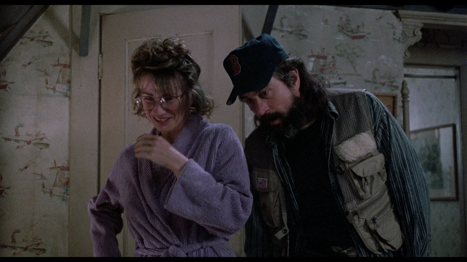 A woman in a purple robe and a man with a beard wearing a baseball cap and a utility vest stand together indoors.