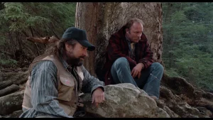 ‘Jacknife’ Blu-Ray Review – Thoughtful Veteran Drama Is A Terrific Acting Showcase Two men sitting in a wooded area near a large tree, one wearing a fishing vest and cap, the other in a plaid jacket.