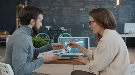 A Practical Approach To Managing Business Processes In One System Two colleagues sitting at a desk and discussing data displayed on a laptop screen.