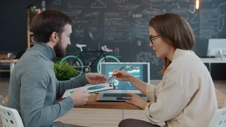 Two colleagues sitting at a desk and discussing data displayed on a laptop screen.